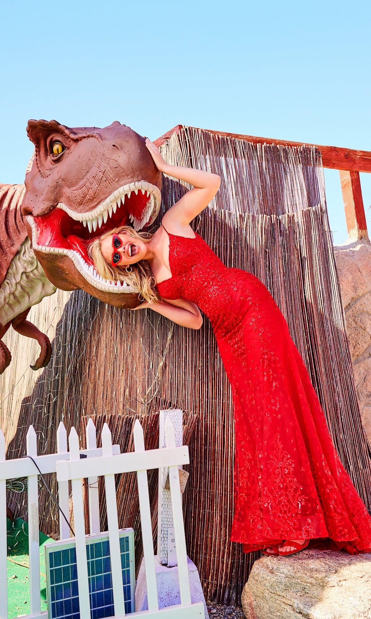 Wearing a Sequin-Lace Open-Back Long Mermaid Prom Dress, a woman playfully poses with her head inside the open mouth of a giant dinosaur statue near a white picket fence.